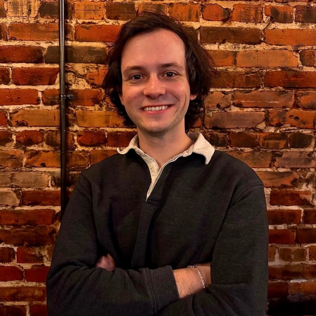 Man standing with arms crossed in front of brick background, smiling.