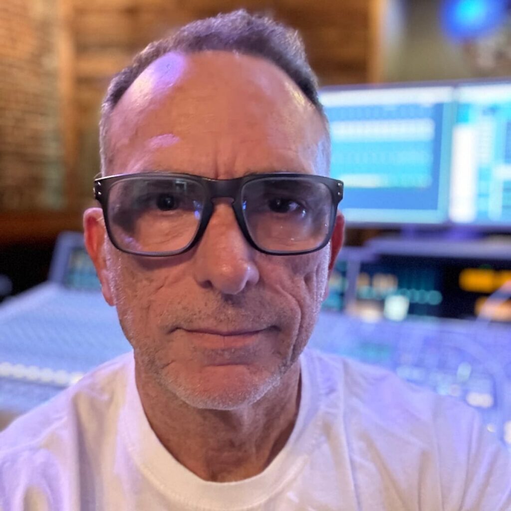 Man with glasses in front of a music recording console.